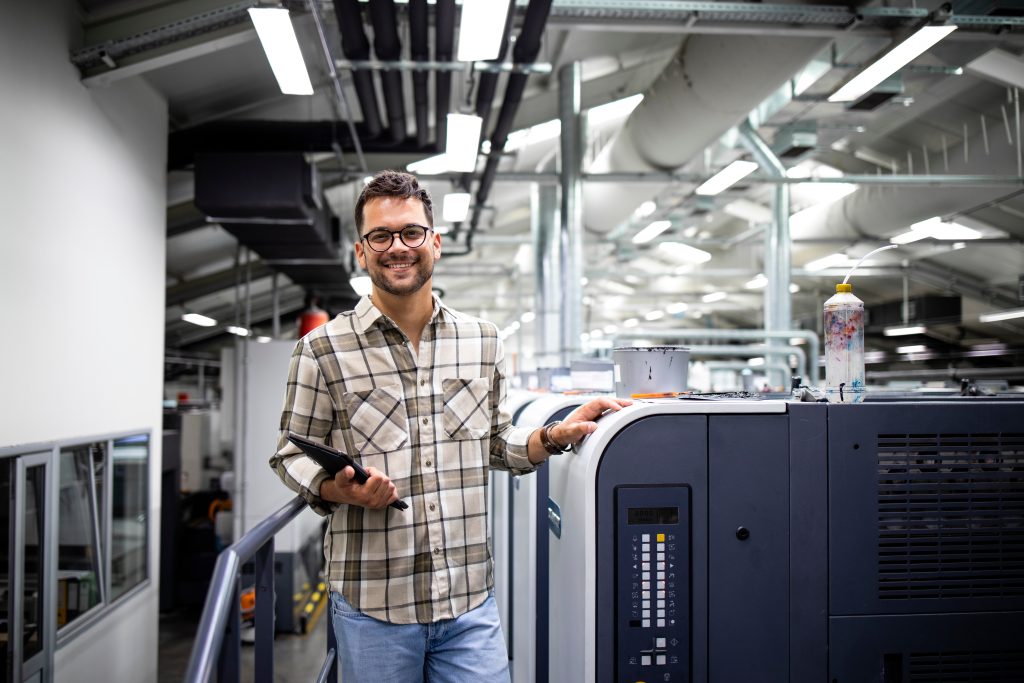 Portrait Of Print Manager Standing By Large Offset Printing Machine In Print Factory.